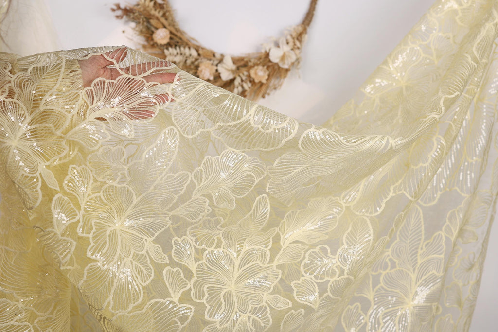 a close-up view of a delicate, yellow fabric with a floral pattern. The fabric appears to be made of sheer material, with a delicate and intricate design. The background is plain white, which allows the fabric to stand out prominently.