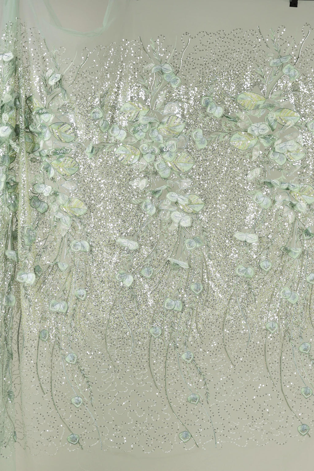 a close-up view of a fabric with a floral pattern, including white and green flowers and leaves. The fabric appears to be made of a shiny, glittery material, giving it a sparkling and delicate appearance.