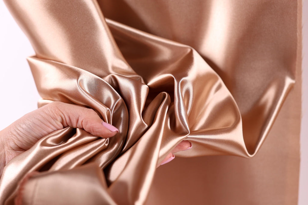 A hand is holding a piece of shiny, golden-colored fabric, which appears to be a satin material.