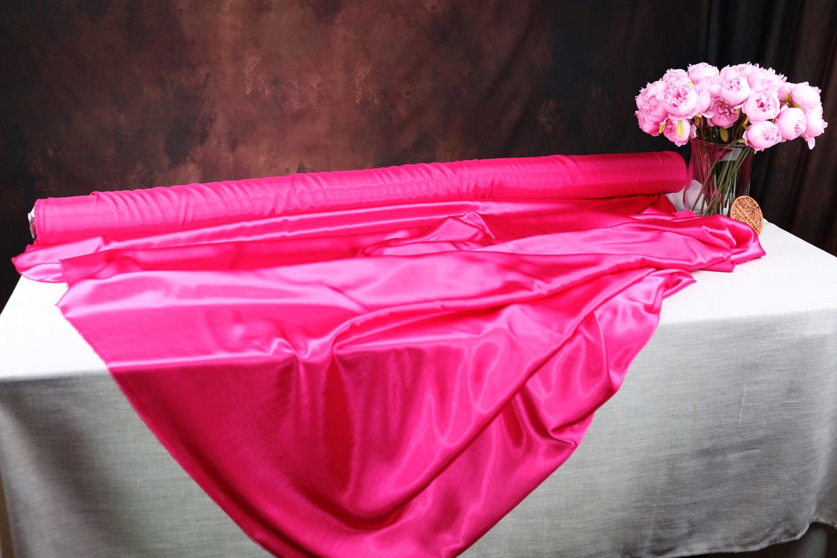 a table with a large roll of shiny pink satin fabric on it, and a vase filled with pink flowers placed next to it.