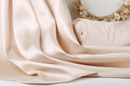 A close-up view of a soft, light pink satin fabric, possibly a sheer or sheer-like material, draped over a surface. In the background, there is a wreath made of dried flowers, adding a decorative element to the scene.