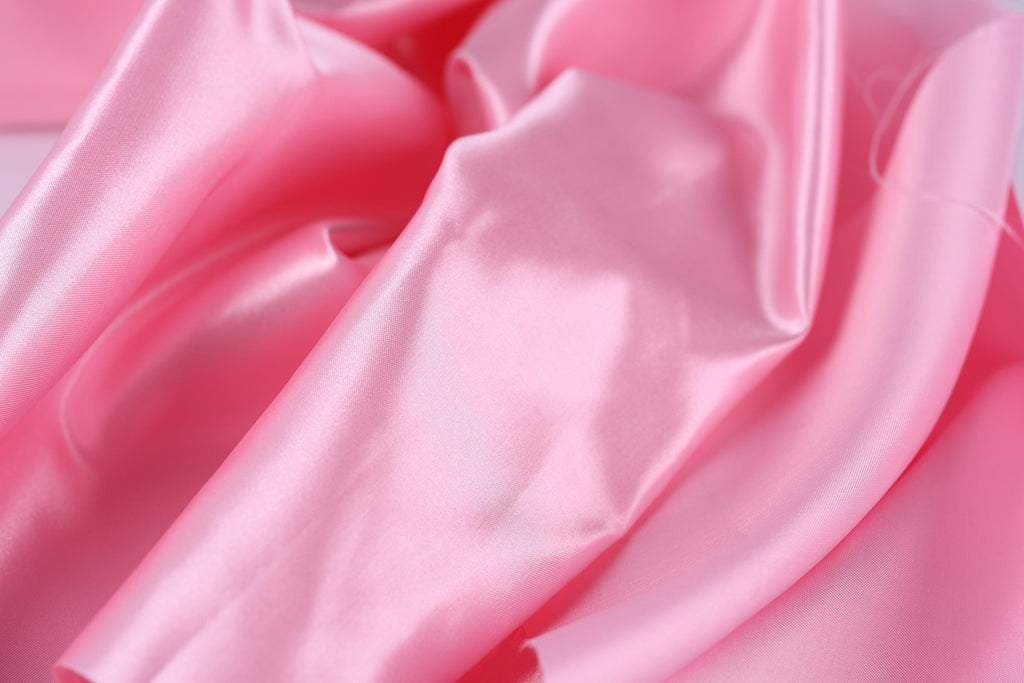 A close-up view of a soft, shiny pink satin fabric, with its folds and creases creating a textured and flowing appearance.