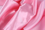 A close-up view of a soft, shiny pink satin fabric, with its folds and creases creating a textured and flowing appearance.