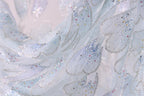 The image is a close-up of a delicate, intricately designed fabric with a light blue color and sparkling embellishments. The fabric appears to be made of sheer material, giving it a soft and delicate appearance. The close-up view allows for a detailed examination of the fabric's texture and patterns, showcasing the craftsmanship and artistry involved in creating such a piece.