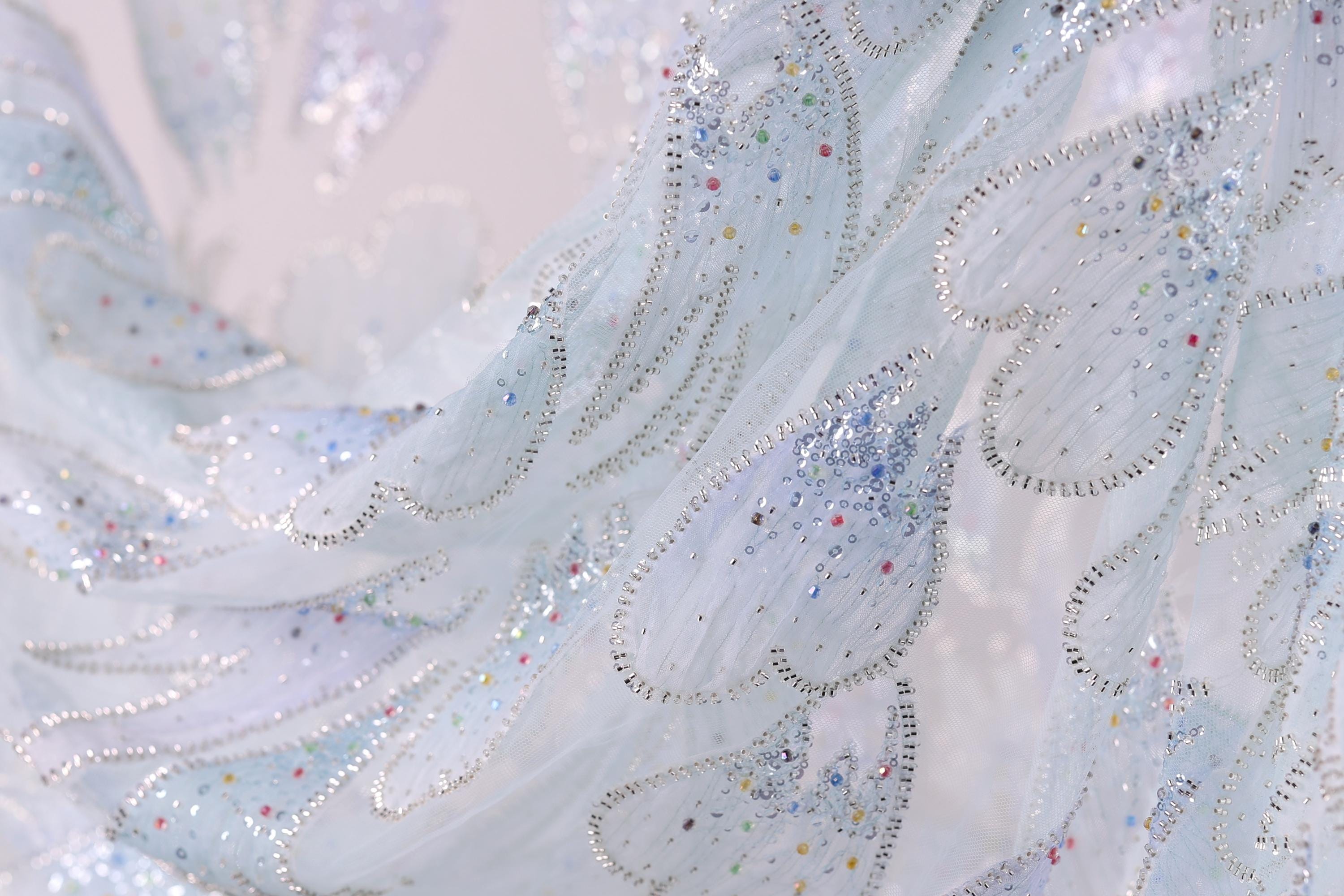 The image is a close-up of a delicate, intricately designed fabric with a light blue color and sparkling embellishments. The fabric appears to be made of sheer material, giving it a soft and delicate appearance. The close-up view allows for a detailed examination of the fabric's texture and patterns, showcasing the craftsmanship and artistry involved in creating such a piece.