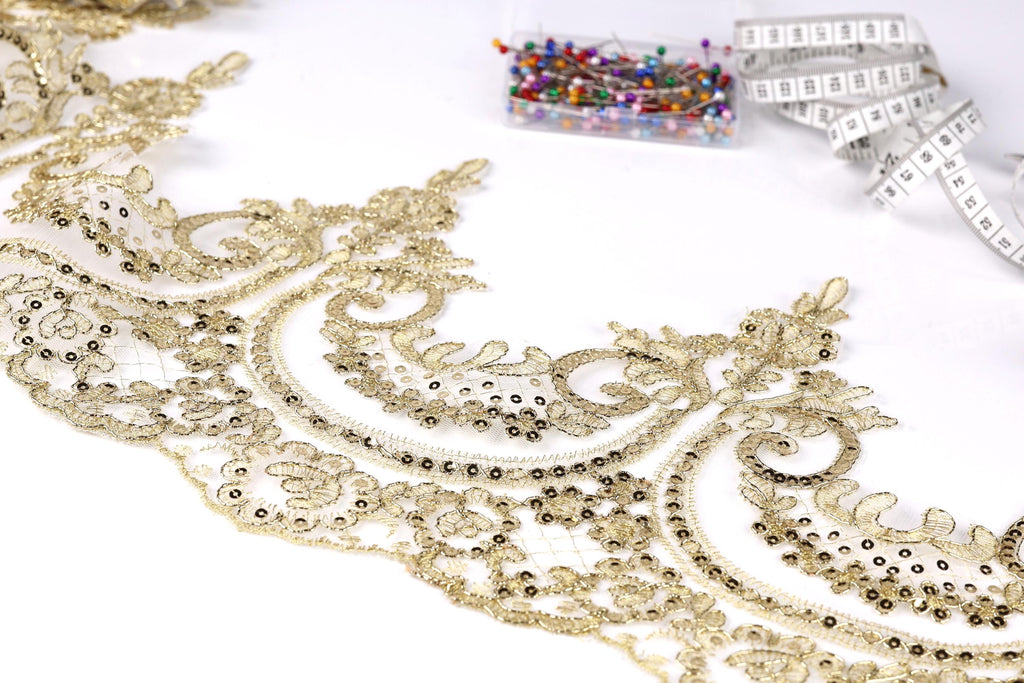a close-up view of a delicate, intricate gold lace fabric with a floral pattern. The fabric appears to be handmade, with a focus on the detailed design and craftsmanship. In the background, there are several small, colorful pins or needles, suggesting that they may be used for sewing or other needlework projects.