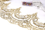 a close-up view of a delicate, intricate gold lace fabric with a floral pattern. The fabric appears to be handmade, with a focus on the detailed design and craftsmanship. In the background, there are several small, colorful pins or needles, suggesting that they may be used for sewing or other needlework projects.