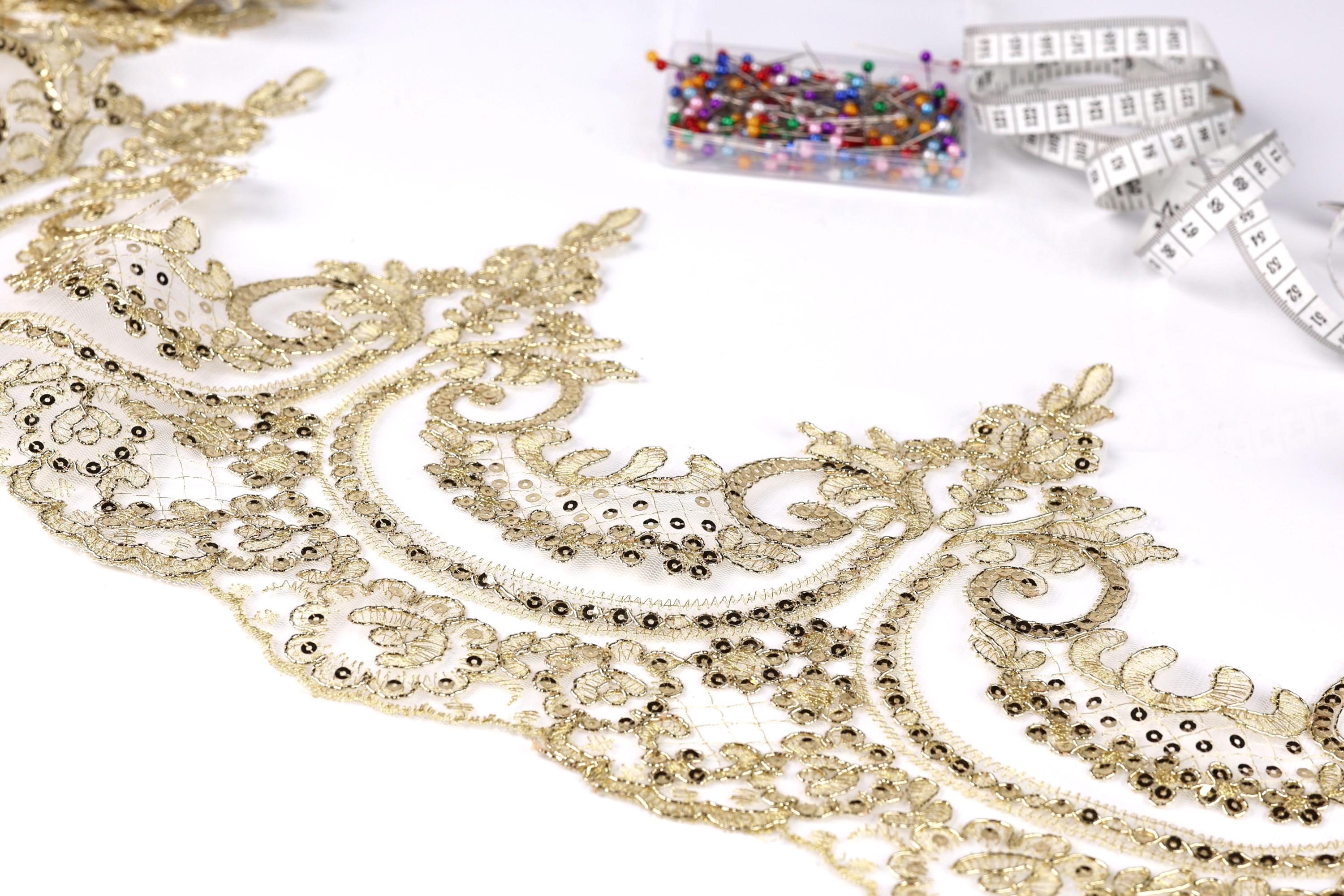 a close-up view of a delicate, intricate gold lace fabric with a floral pattern. The fabric appears to be handmade, with a focus on the detailed design and craftsmanship. In the background, there are several small, colorful pins or needles, suggesting that they may be used for sewing or other needlework projects.