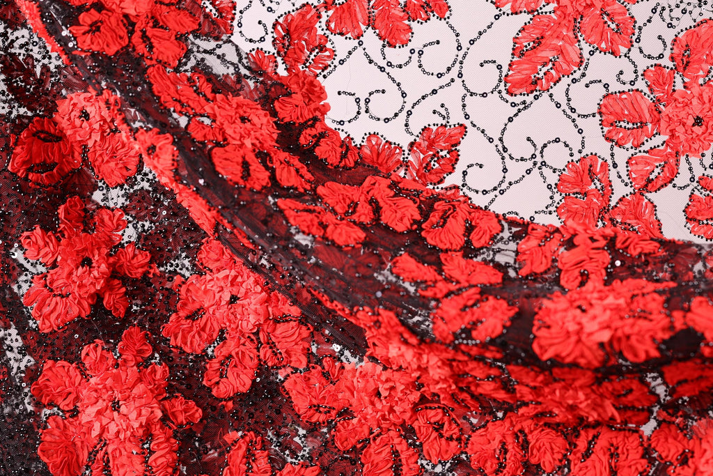The image is a close-up of a fabric with a striking red and black floral pattern. The fabric appears to be made of lace or a similar material, with a repeating floral design in shades of red and black. The fabric is intricately detailed, showcasing the craftsmanship and artistry involved in its creation.