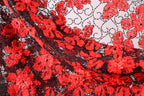 The image is a close-up of a fabric with a striking red and black floral pattern. The fabric appears to be made of lace or a similar material, with a repeating floral design in shades of red and black. The fabric is intricately detailed, showcasing the craftsmanship and artistry involved in its creation.