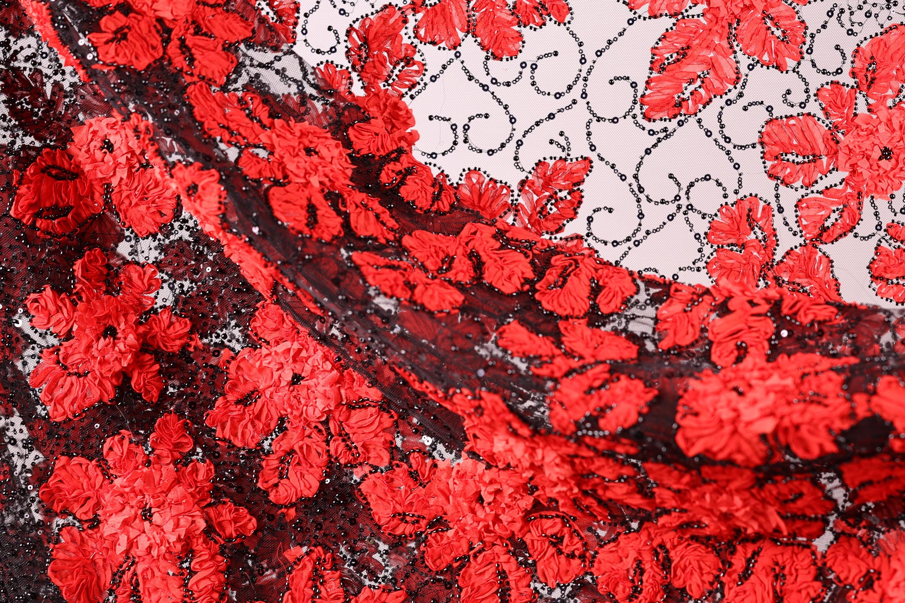 The image is a close-up of a fabric with a striking red and black floral pattern. The fabric appears to be made of lace or a similar material, with a repeating floral design in shades of red and black. The fabric is intricately detailed, showcasing the craftsmanship and artistry involved in its creation.
