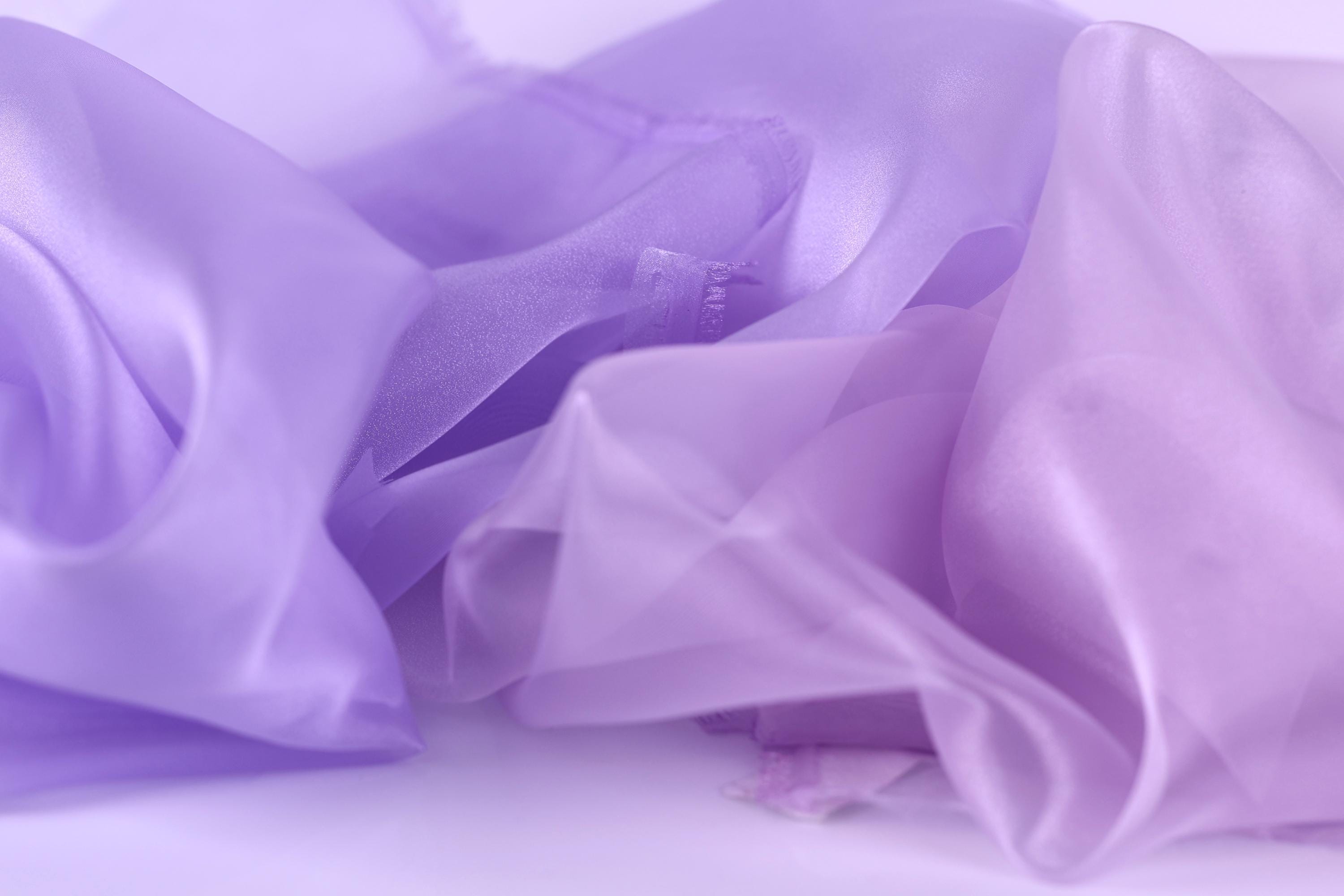 The image is a close-up of a soft, flowing fabric in shades of purple and pink, with a slightly crumpled texture.
