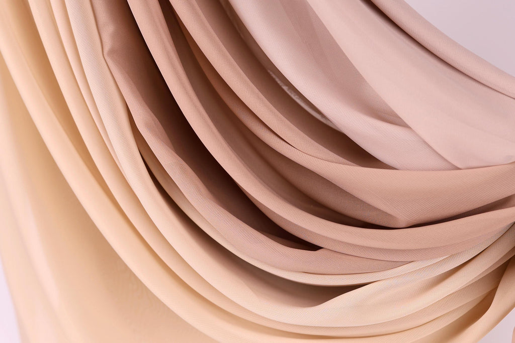 a close-up view of a fabric with a gradient of colors, transitioning from a light beige to a darker beige. The fabric appears to be folded or draped in a way that creates a sense of movement and texture.