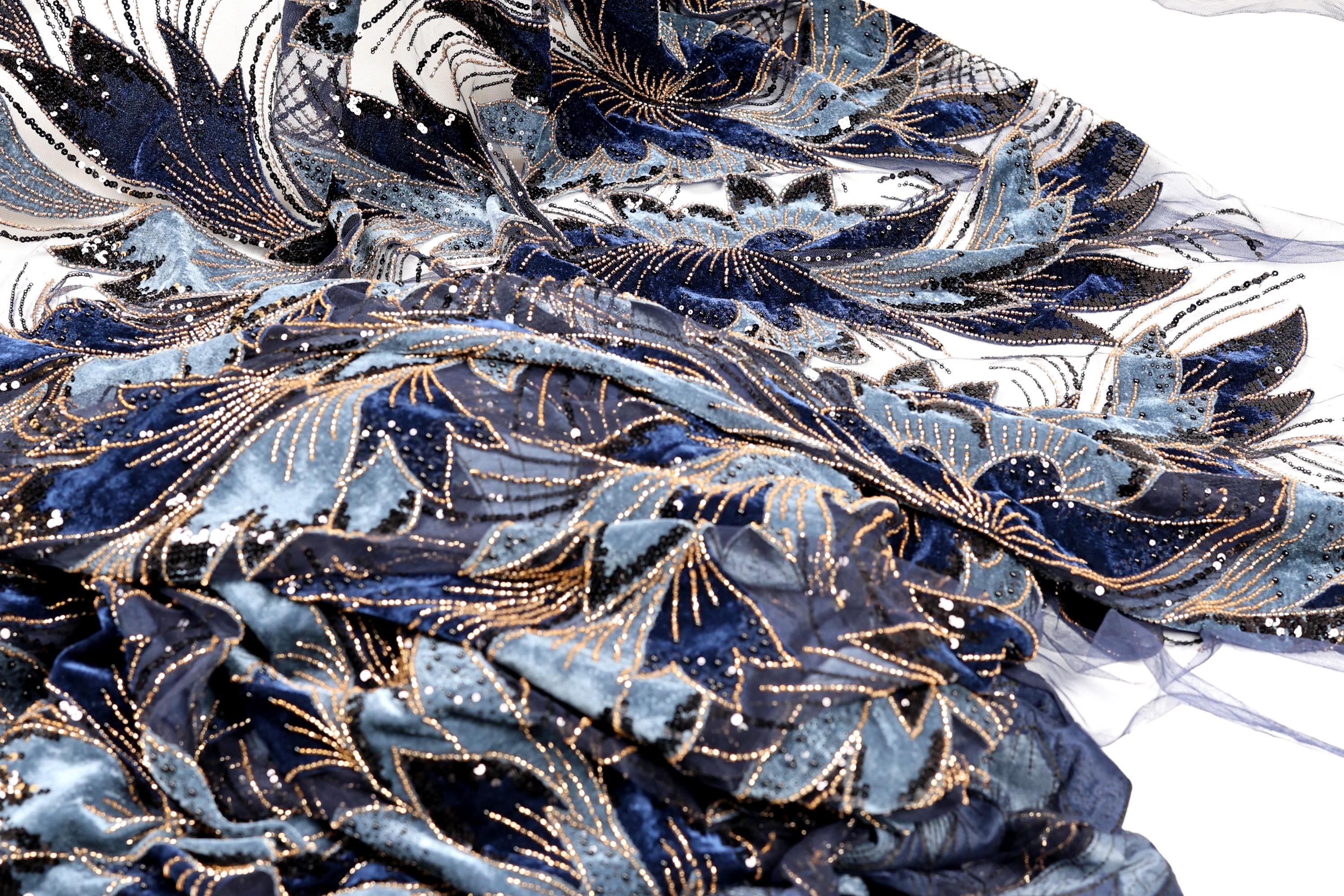 a close-up view of a fabric with a blue and gold floral pattern. The fabric appears to be made of a shiny material, possibly sequin or lace, and has a textured, crumpled appearance.