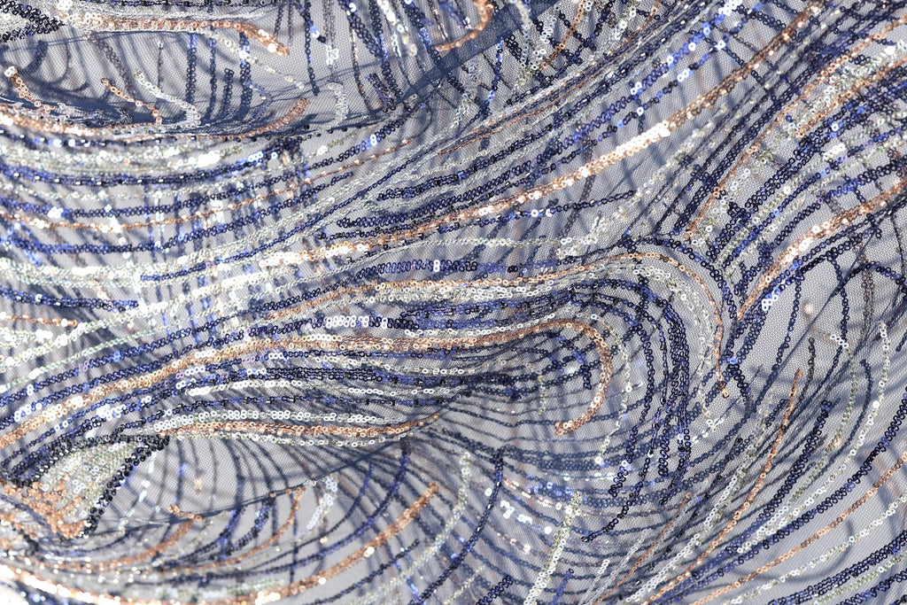 The image is a close-up view of a fabric with a complex, swirling pattern of blue, silver, and white sequins. The fabric appears to be made of a shiny, metallic material, giving it a metallic, glittery appearance. The pattern is intricate and visually striking, with the sequins creating a sense of movement and depth.