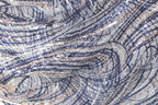 The image is a close-up view of a fabric with a complex, swirling pattern of blue, silver, and white sequins. The fabric appears to be made of a shiny, metallic material, giving it a metallic, glittery appearance. The pattern is intricate and visually striking, with the sequins creating a sense of movement and depth.