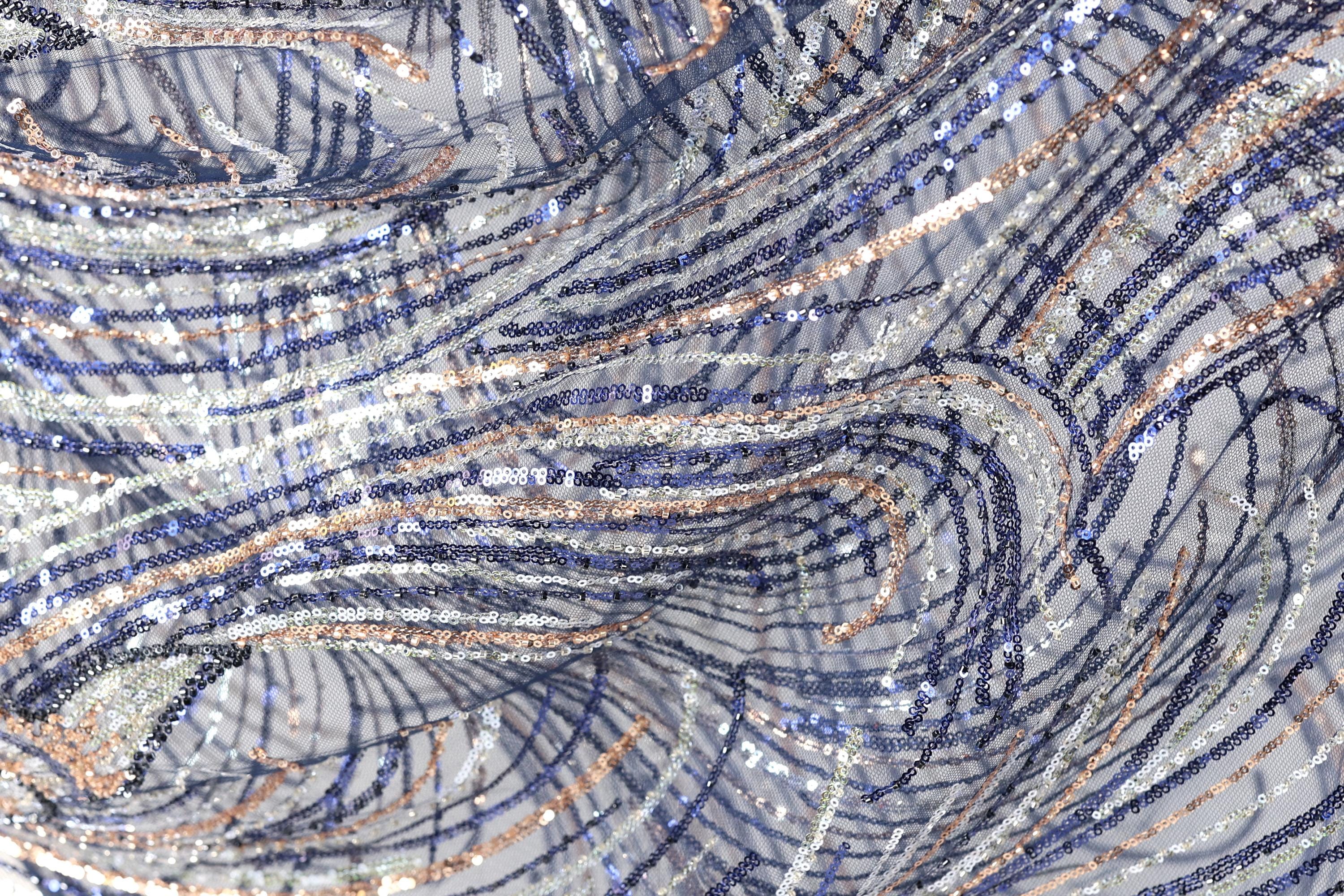 The image is a close-up view of a fabric with a complex, swirling pattern of blue, silver, and white sequins. The fabric appears to be made of a shiny, metallic material, giving it a metallic, glittery appearance. The pattern is intricate and visually striking, with the sequins creating a sense of movement and depth.