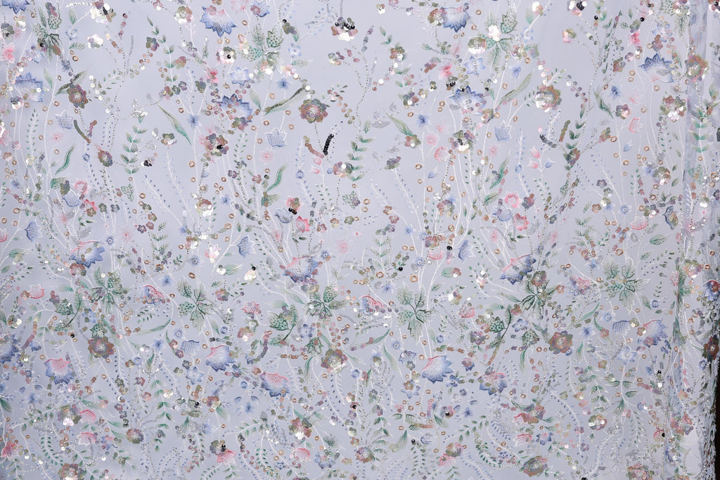 The image is a close-up of a fabric with a floral pattern, featuring a variety of colorful flowers and leaves. The fabric appears to be made of a light-colored material, possibly lace or a similar type of textile. The pattern is intricate and detailed, with each flower and leaf meticulously crafted to create a visually appealing and textured design.