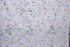 The image is a close-up of a fabric with a floral pattern, featuring a variety of colorful flowers and leaves. The fabric appears to be made of a light-colored material, possibly lace or a similar type of textile. The pattern is intricate and detailed, with each flower and leaf meticulously crafted to create a visually appealing and textured design.