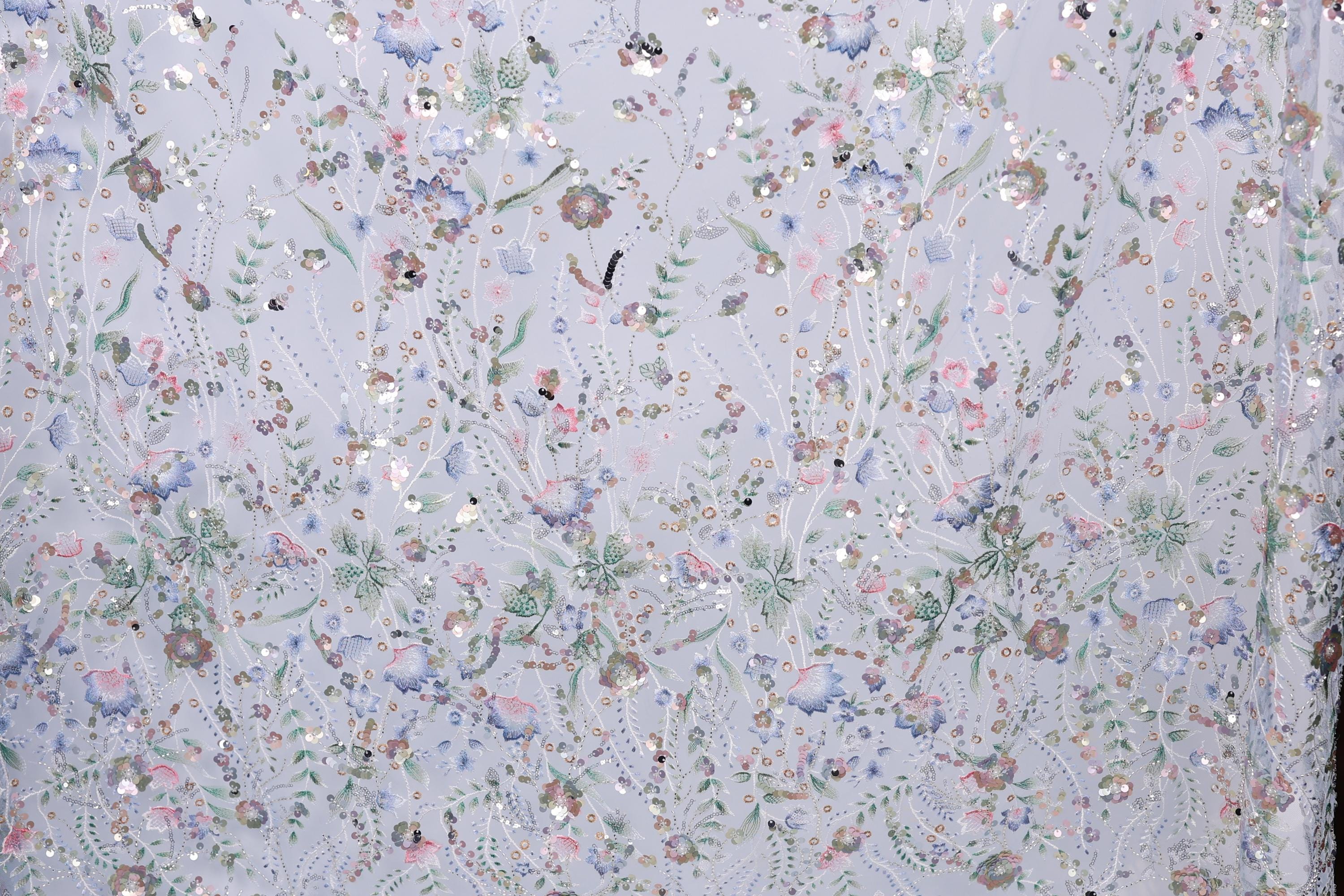 The image is a close-up of a fabric with a floral pattern, featuring a variety of colorful flowers and leaves. The fabric appears to be made of a light-colored material, possibly lace or a similar type of textile. The pattern is intricate and detailed, with each flower and leaf meticulously crafted to create a visually appealing and textured design.