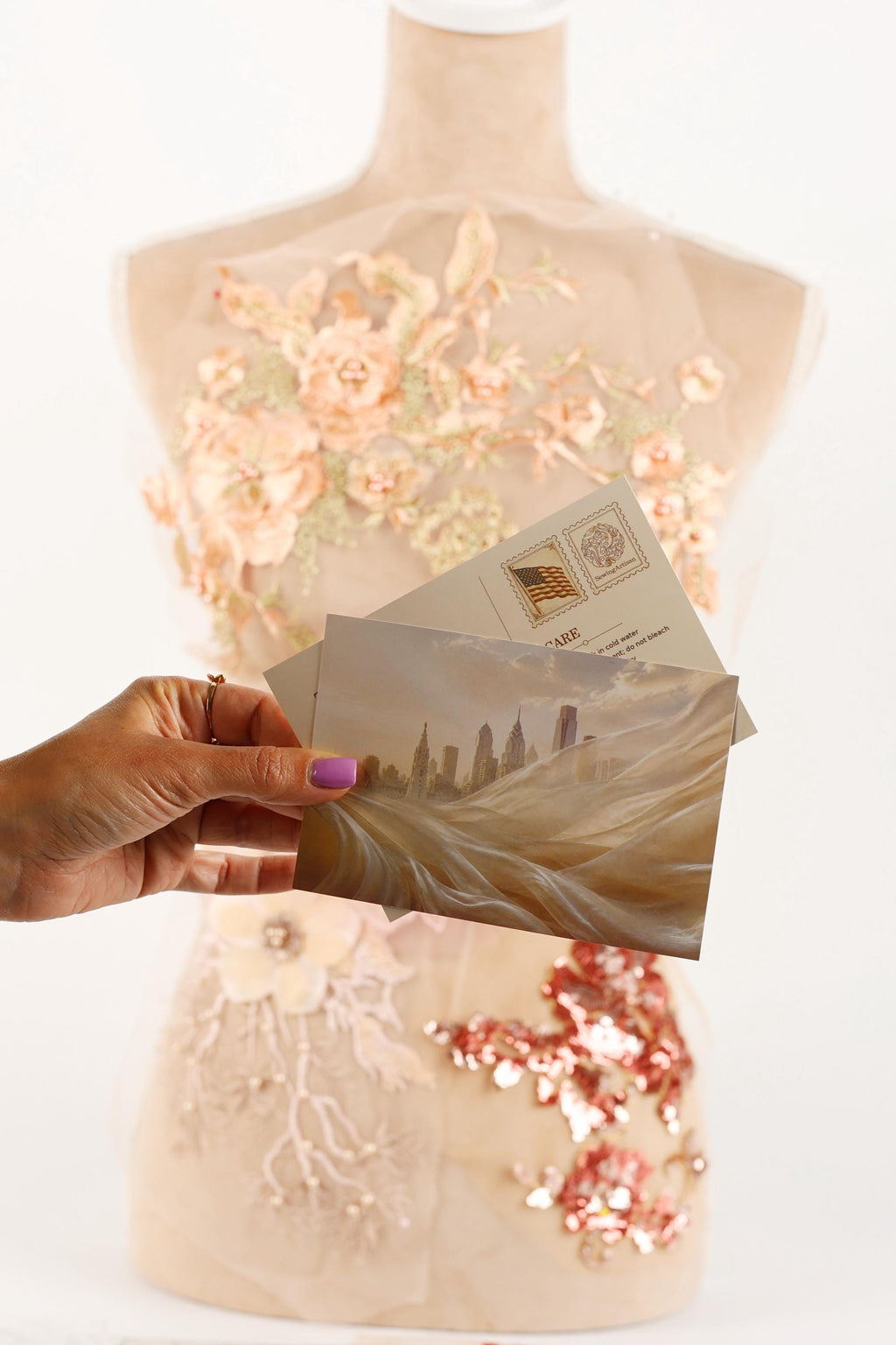 A hand holding a postcard with a cityscape image, against a background of a mannequin with a floral dress and sequin embellishments.