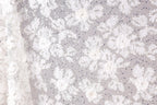 The image is a close-up of a delicate, white lace fabric with a floral pattern. The fabric appears to be made of a sheer material, giving it a delicate and elegant appearance. The lace is adorned with small white flowers and beads, adding a touch of sparkle and detail to the overall design. The image does not contain any text or other discernible objects, and the focus is solely on the intricate lace pattern.