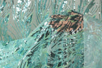 a close-up view of a fabric with a floral pattern and sequin details. The fabric appears to be a light blue color, with a darker blue and green pattern visible. The background is a light gray color, providing a neutral backdrop for the fabric.