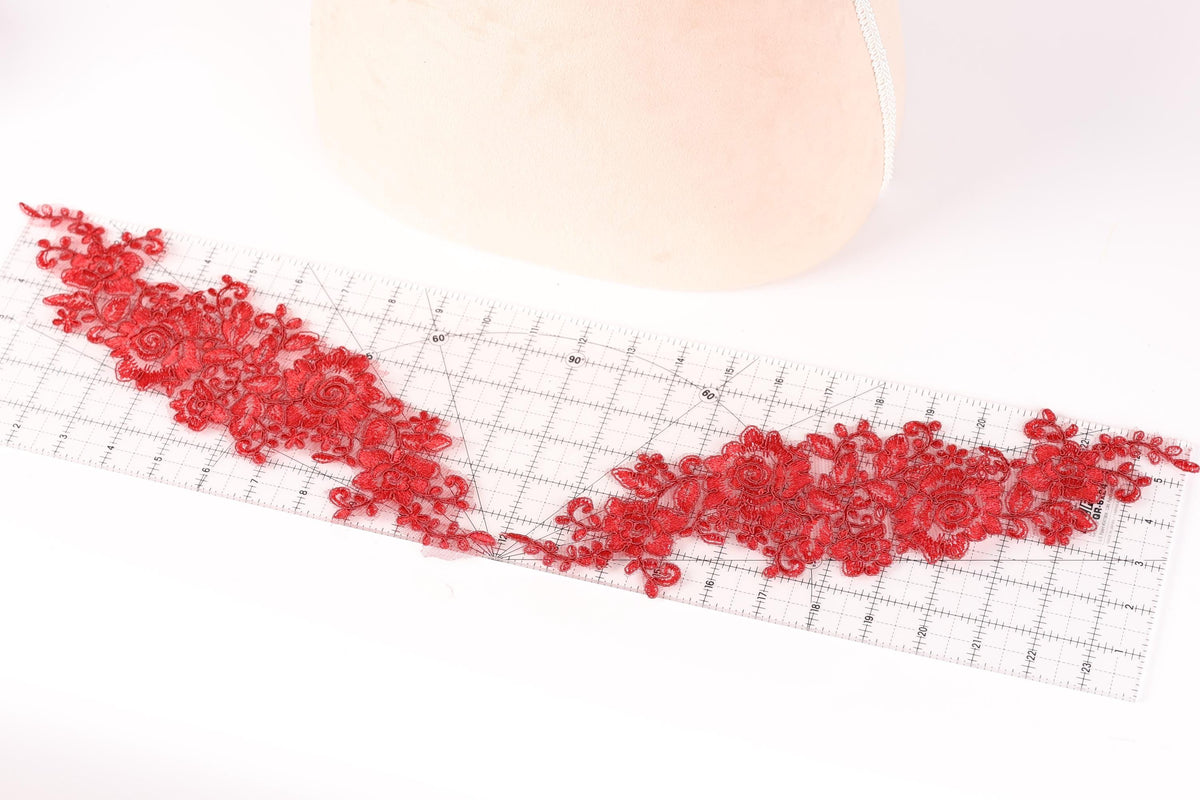 a piece of red lace with intricate floral patterns laid out on a transparent ruler, which is placed on a white surface.