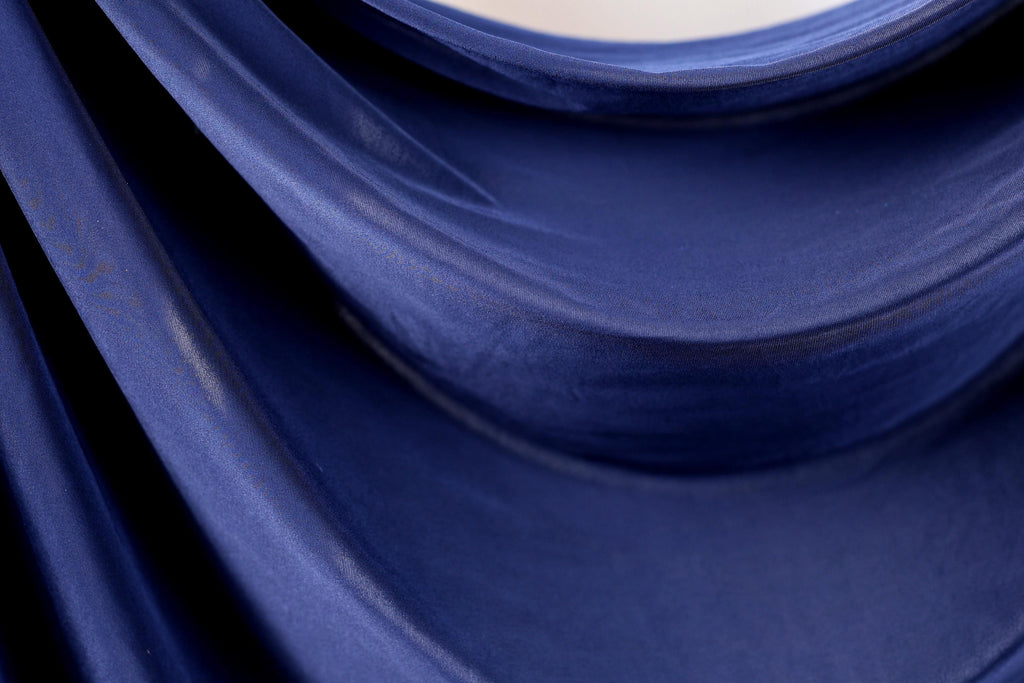 a close-up view of a blue fabric, possibly a piece of clothing or a textile. The fabric appears to be folded or draped, creating a sense of movement and texture. The color of the fabric is dark blue, which contrasts with the lighter background. The image does not contain any discernible objects or actions, and there are no specific actions or movements visible. The focus is on the fabric and its texture, with no additional context provided.