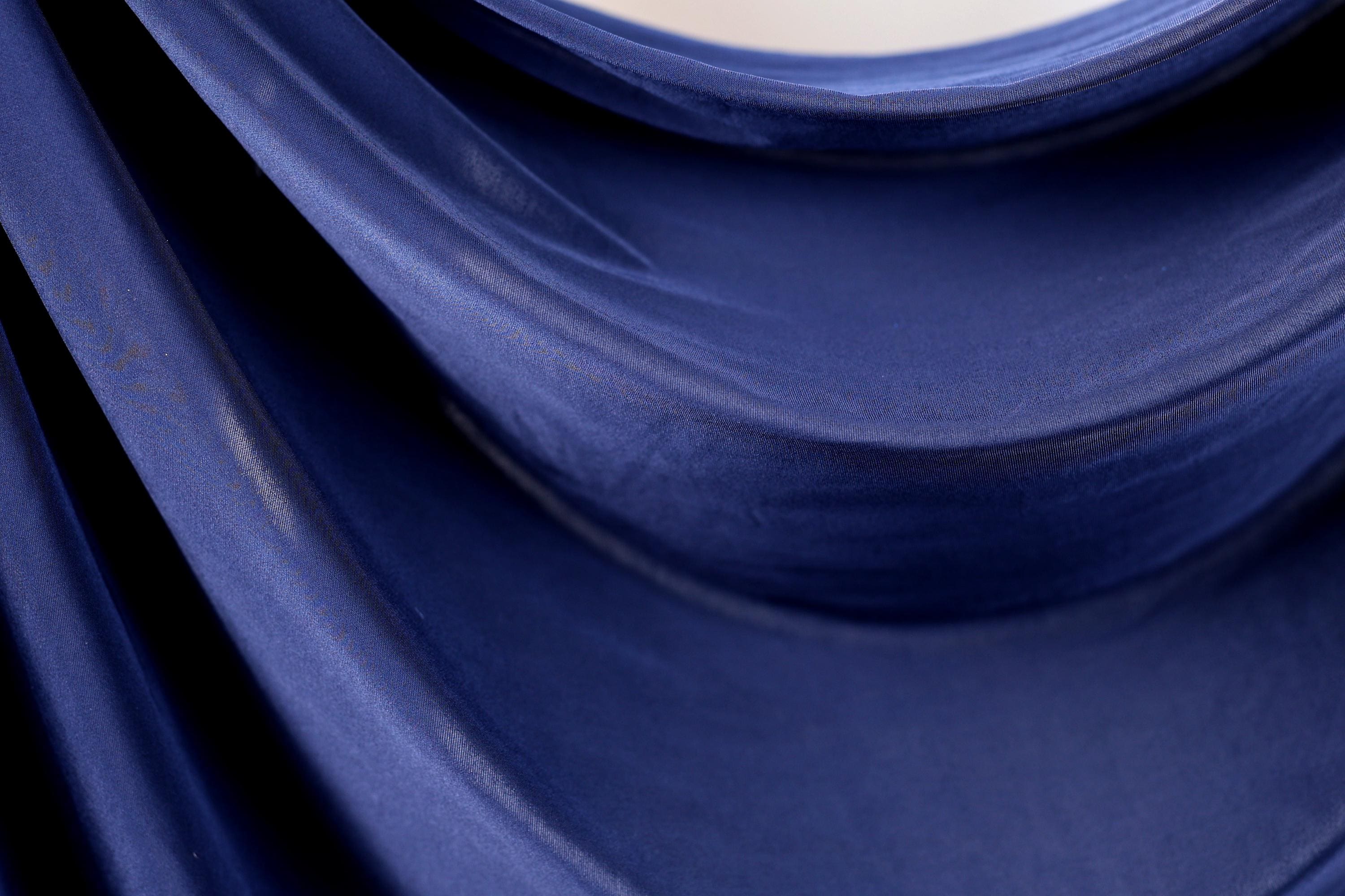 a close-up view of a blue fabric, possibly a piece of clothing or a textile. The fabric appears to be folded or draped, creating a sense of movement and texture. The color of the fabric is dark blue, which contrasts with the lighter background. The image does not contain any discernible objects or actions, and there are no specific actions or movements visible. The focus is on the fabric and its texture, with no additional context provided.