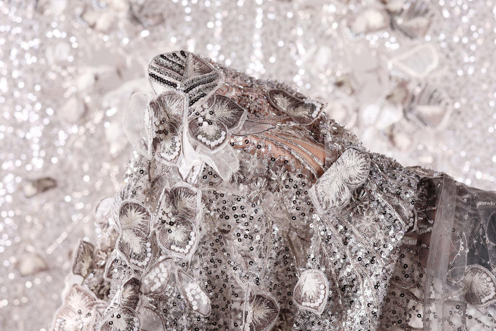 a close-up view of a delicate, intricately designed fabric with a floral pattern. The fabric appears to be made of sequins and lace, creating a visually striking and elegant appearance.