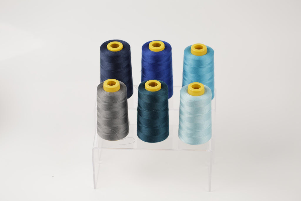A collection of six spools of thread in various colors, including blue, gray, and light blue, arranged on a clear stand.