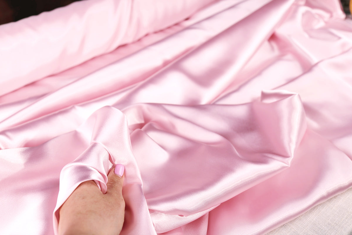 A hand is holding a piece of pink satin fabric, which appears to be a soft and smooth material.
