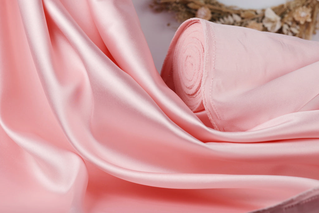 A close-up view of a soft, flowing pink satin fabric, with a rolled-up piece of the fabric visible in the background.
