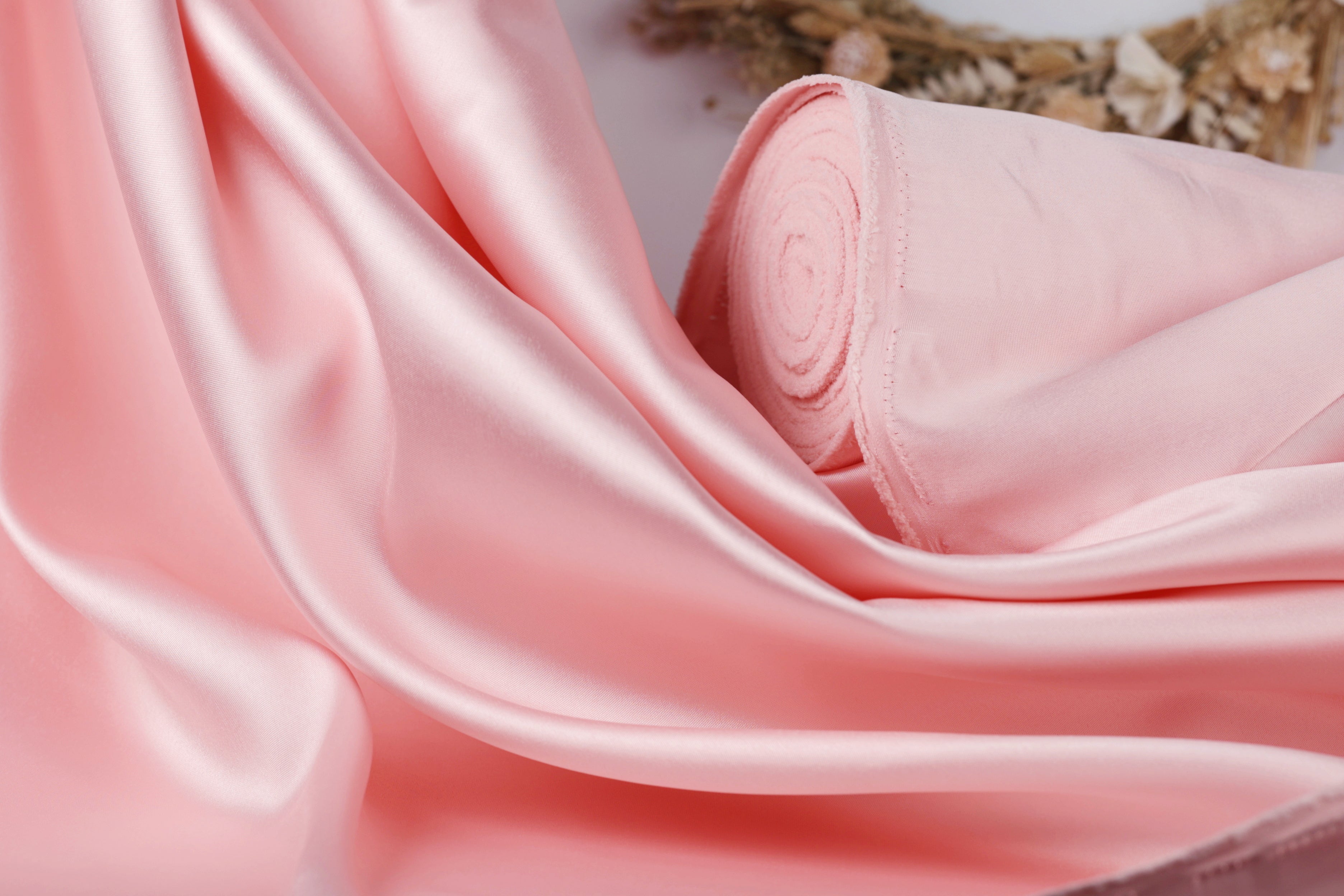 A close-up view of a soft, flowing pink satin fabric, with a rolled-up piece of the fabric visible in the background.
