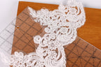 a close-up view of a delicate, intricate white lace fabric with floral patterns. The lace appears to be a part of a larger piece of fabric, as it is placed on a wooden surface. There are also two rulers visible in the image, one of which is partially visible on the left side.