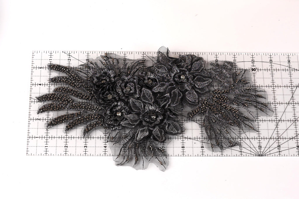 a close-up view of a black lace or fabric with intricate floral or leaf-like patterns. The lace appears to be made of a shiny material, possibly beads or sequins, and is displayed on a ruler for scale.