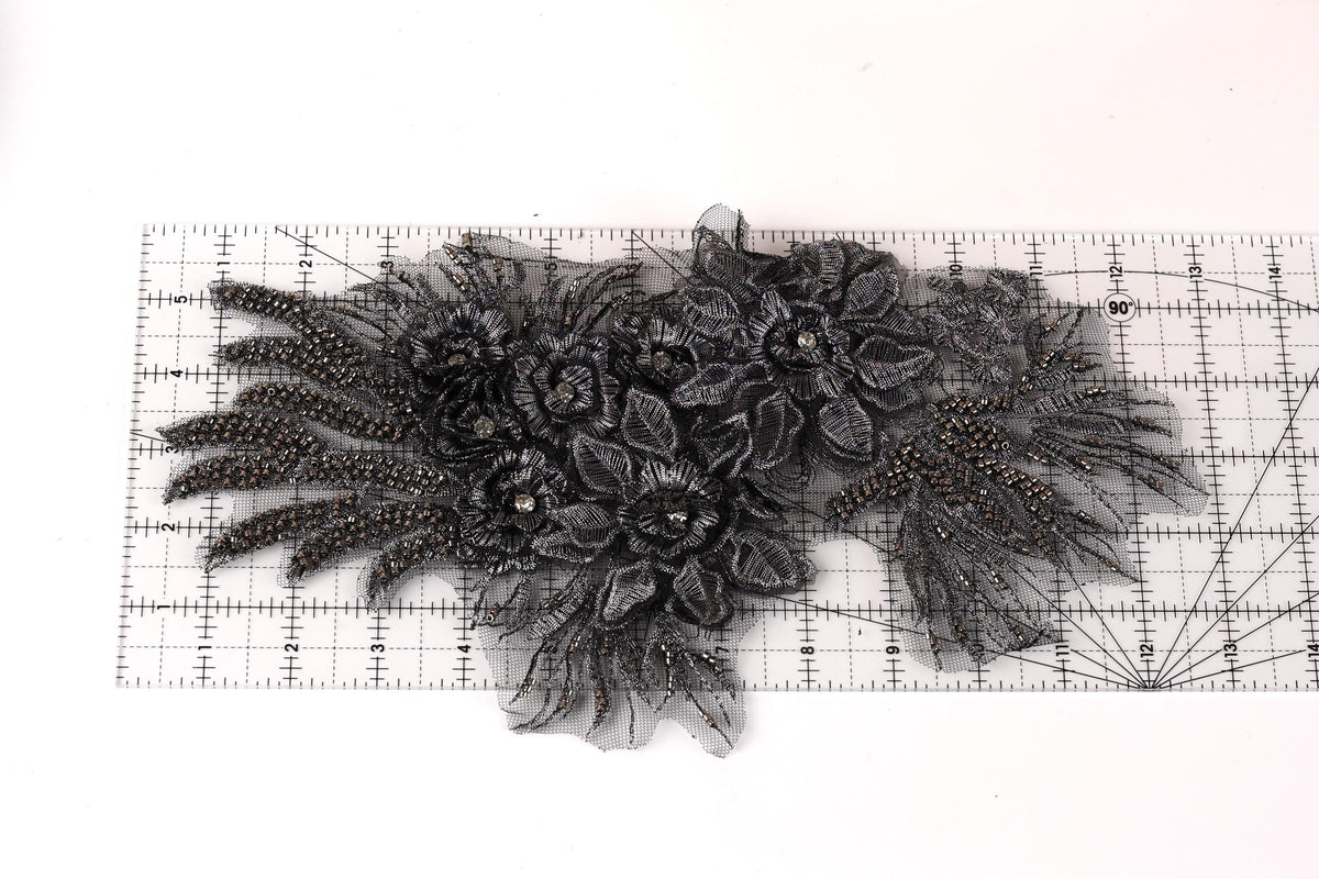 a close-up view of a black lace or fabric with intricate floral or leaf-like patterns. The lace appears to be made of a shiny material, possibly beads or sequins, and is displayed on a ruler for scale.