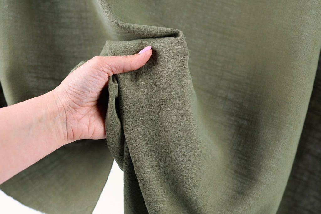 A hand is holding a piece of olive green fabric, which appears to be a type of fabric or textile.