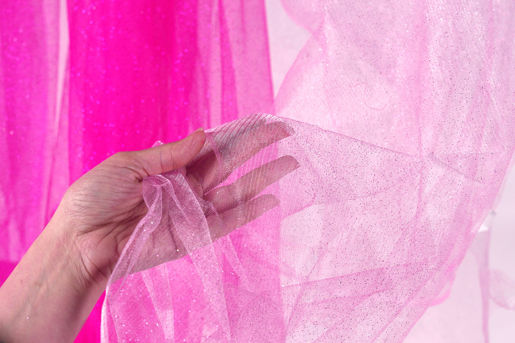 A hand is holding a piece of pink glittery fabric against a pink background.