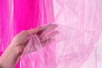 A hand is holding a piece of pink glittery fabric against a pink background.