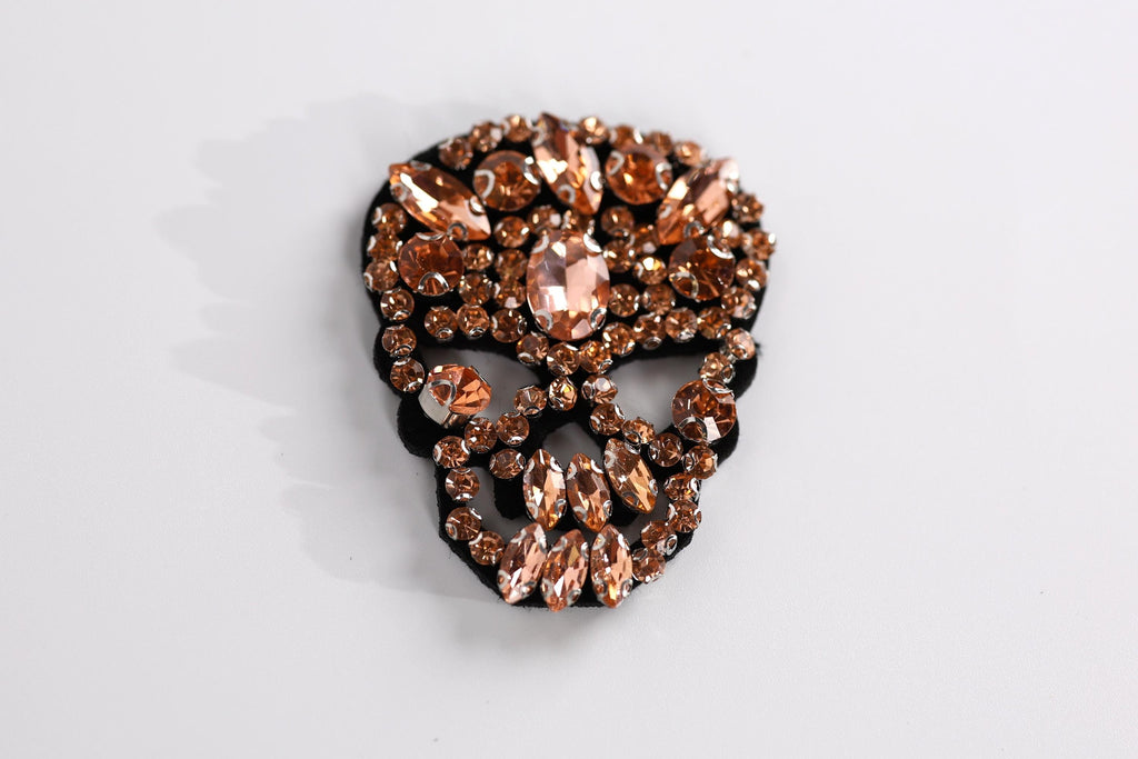 A decorative skull-shaped brooch adorned with numerous small, clear gemstones in shades of brown and orange.