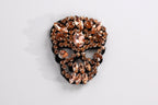 A decorative skull-shaped brooch adorned with numerous small, clear gemstones in shades of brown and orange.