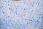 a close-up view of a fabric with a floral pattern of blue flowers and leaves. The fabric appears to be made of a light-colored material, possibly cotton or linen, and has a delicate, intricate design. The pattern consists of numerous small blue flowers and leaves, arranged in a repeating pattern across the fabric. The overall appearance of the fabric is serene and elegant, with the blue flowers and leaves adding a touch of natural beauty to the design.