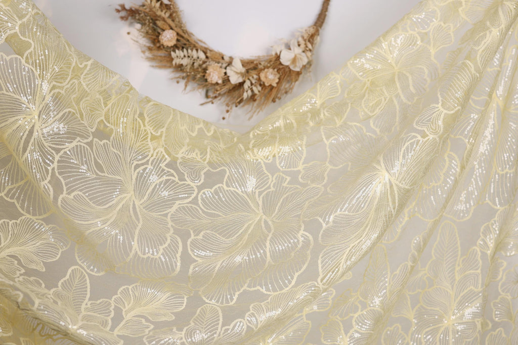 a close-up view of a delicate, yellow fabric with a floral pattern. The fabric appears to be made of sheer material, with a delicate and intricate design. The background is plain white, allowing the fabric and floral pattern to stand out prominently.