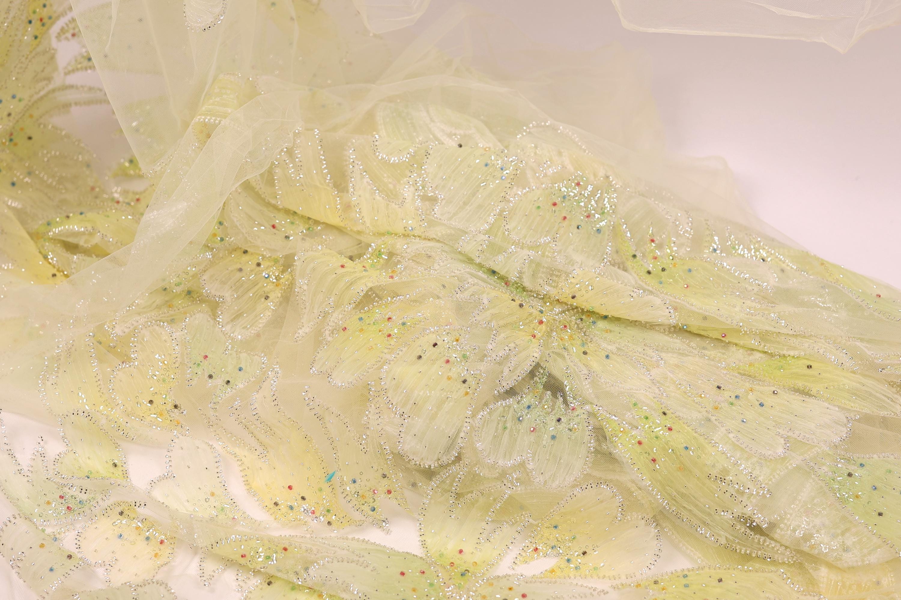 a close-up view of a delicate, sheer fabric with a yellow floral pattern. The fabric appears to be made of sheer material, giving it a soft and delicate appearance. The pattern consists of small yellow flowers or leaves, which are intricately designed and embellished with small, colorful dots or sequins. The fabric is spread out, revealing its intricate details and textures, creating a visually appealing and delicate piece of textile art.