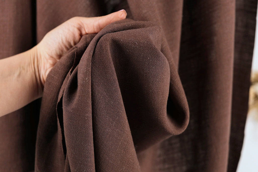 A hand is holding a brown fabric, possibly a piece of clothing or a textile, in front of a brown curtain.