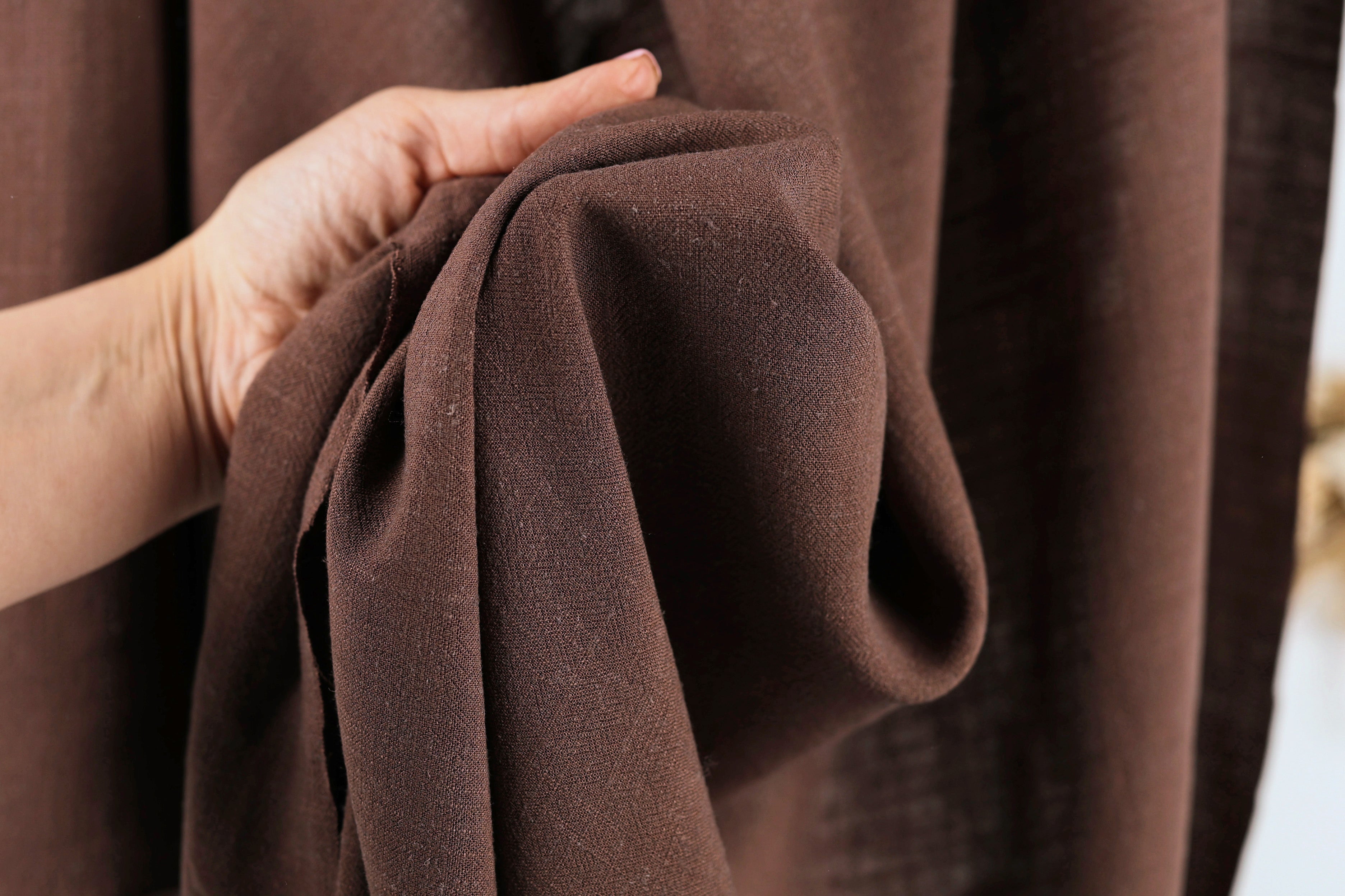 A hand is holding a brown fabric, possibly a piece of clothing or a textile, in front of a brown curtain.