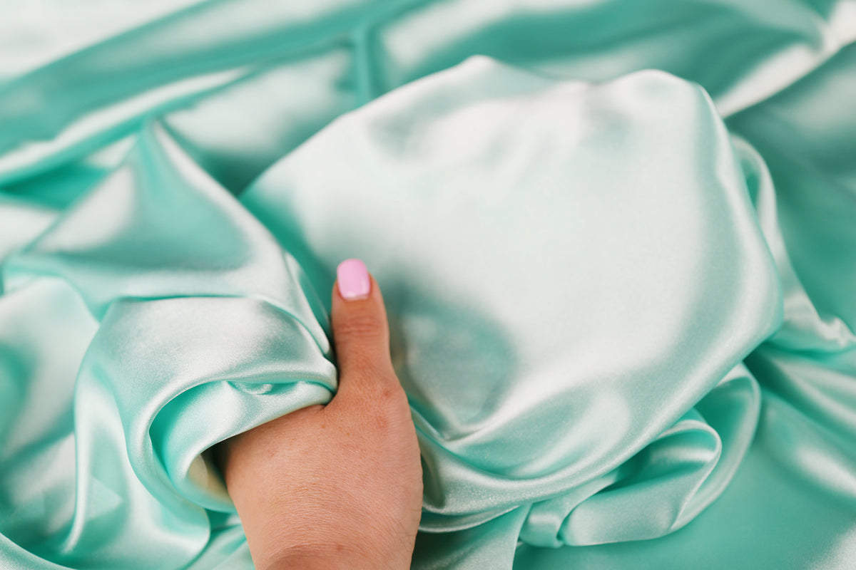 A hand with pink nail polish is holding a piece of light blue satin fabric.