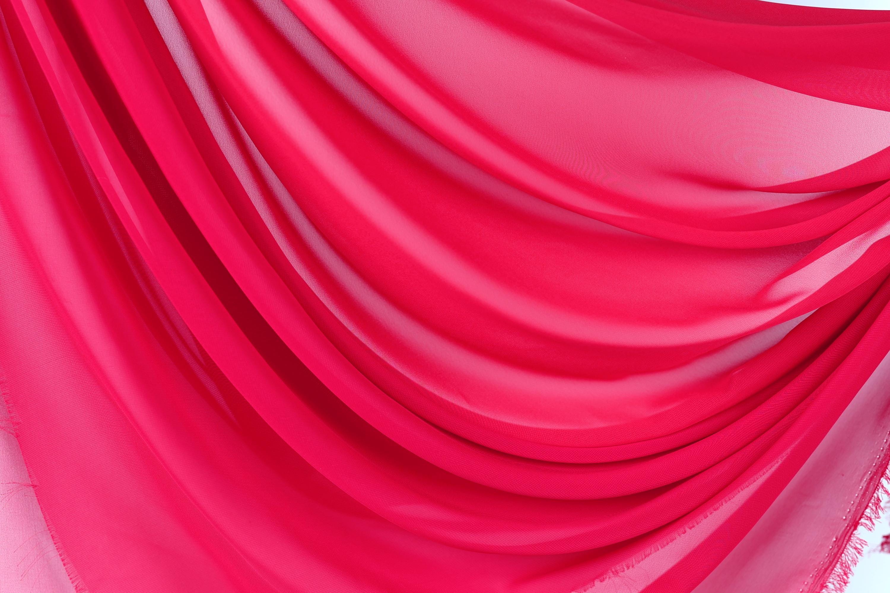 The image is a close-up of a vibrant red fabric, possibly a chiffon or sheer material, with a smooth, flowing texture. The fabric appears to be draped or draped over an object, creating a sense of movement and depth. The color and texture of the fabric are the main focus of the image, with no other objects or details visible.